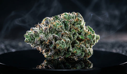A macro shot of a frost-covered cannabis bud on a black reflective surface with subtle smoke.